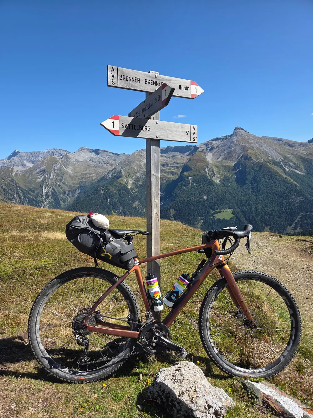 Das Gravelbike mit Satteltasche lehnt an einem Wegweiser Richtung Brenner, dahinter Berge unter blauem Himmel