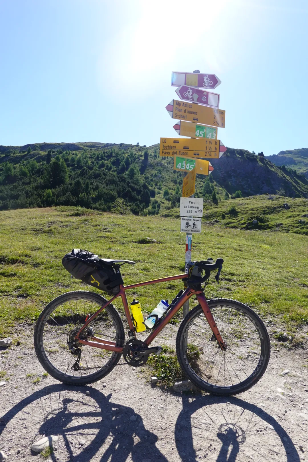 Gravelbike unter einem Wegweiser auf einer Alpwiese in der Morgensonne