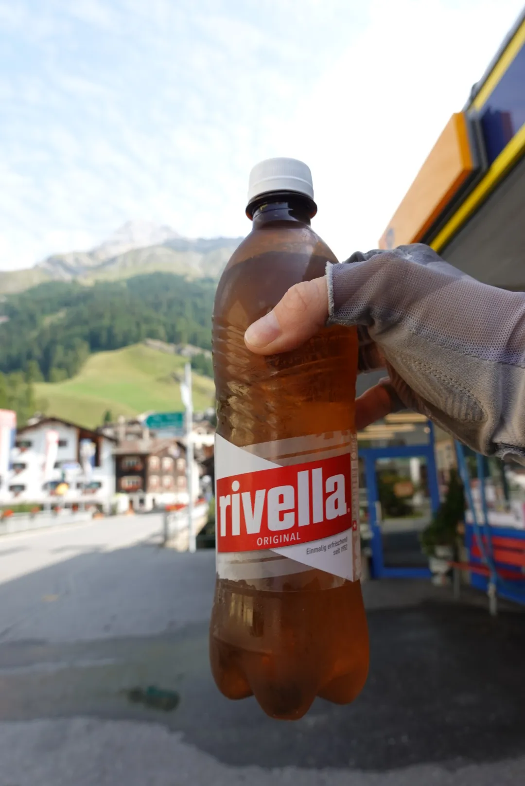 Eine Flasche Rivella in der Hand, dahinter ein Bergdorf
