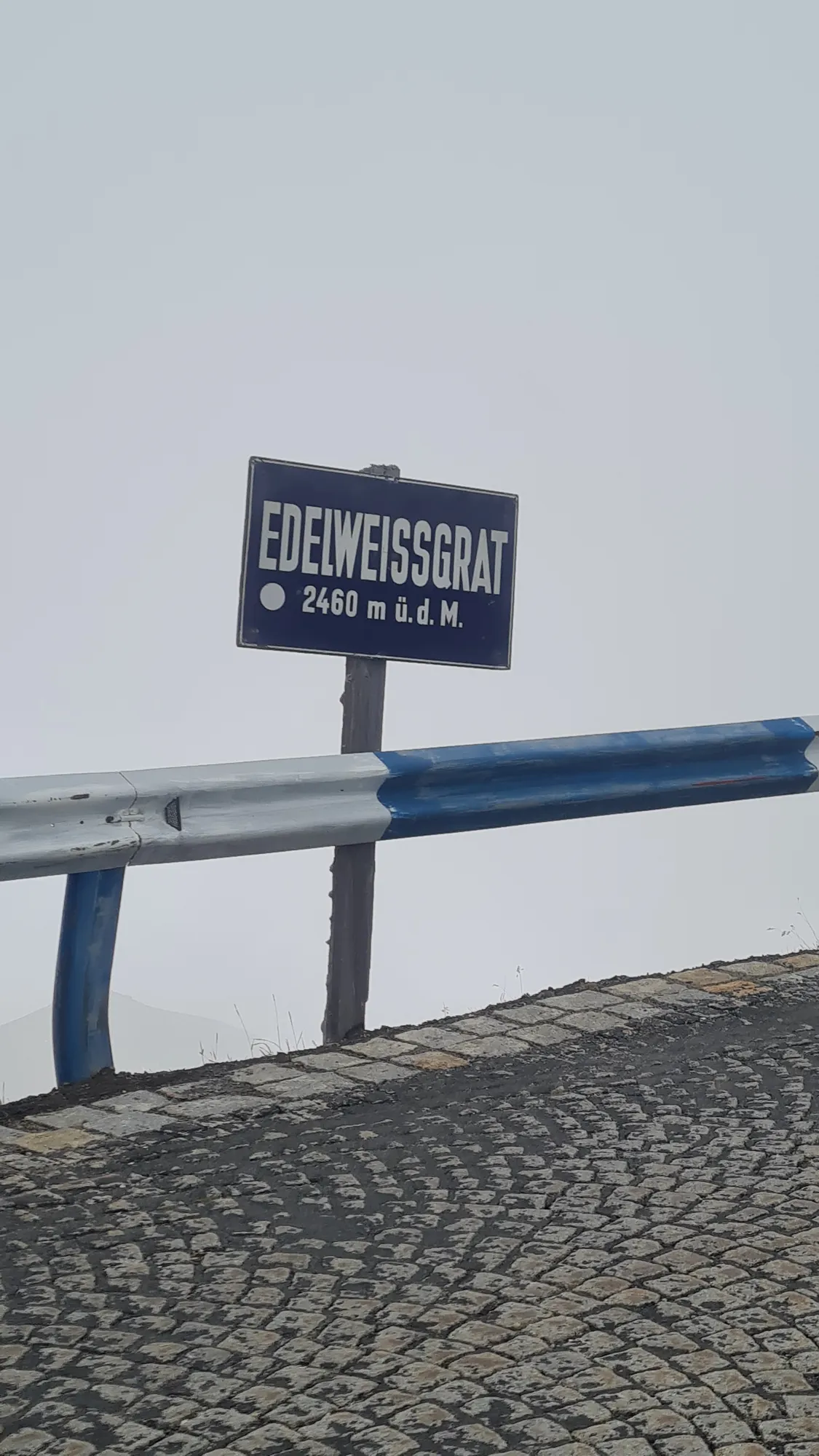 Schild „Edelweissgrat“ an der gepflasterten Straße im Nebel