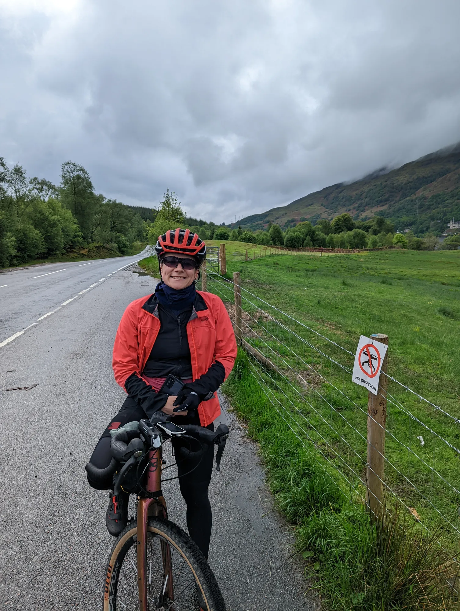 Carla in roter Jacke mit dem Rad am Straßenrand, dahinter grüne Hügel