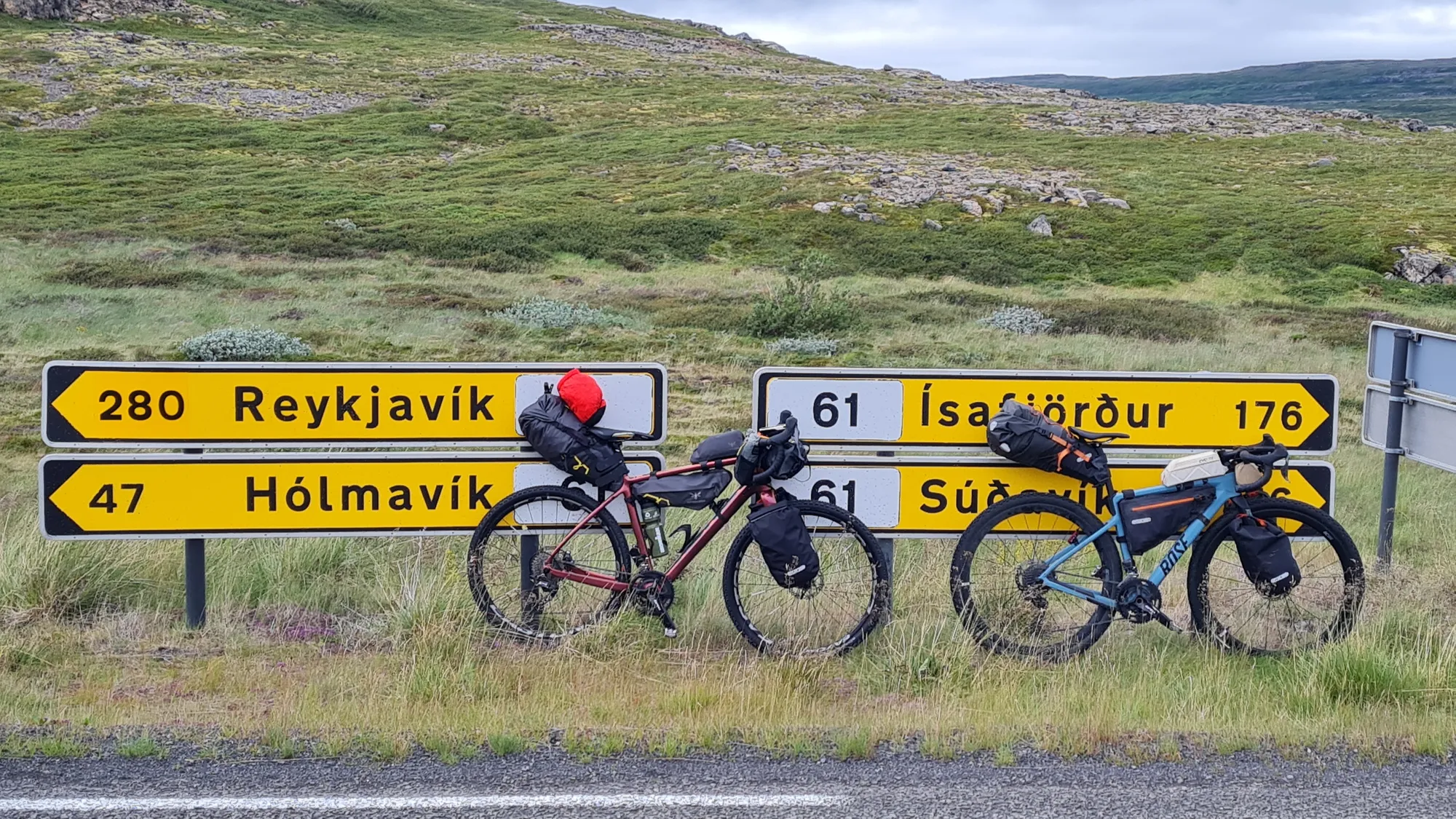 Zwei bepackte Gravelbikes vor gelben Wegweisern nach Reykjavík, Hólmavík und Ísafjörður